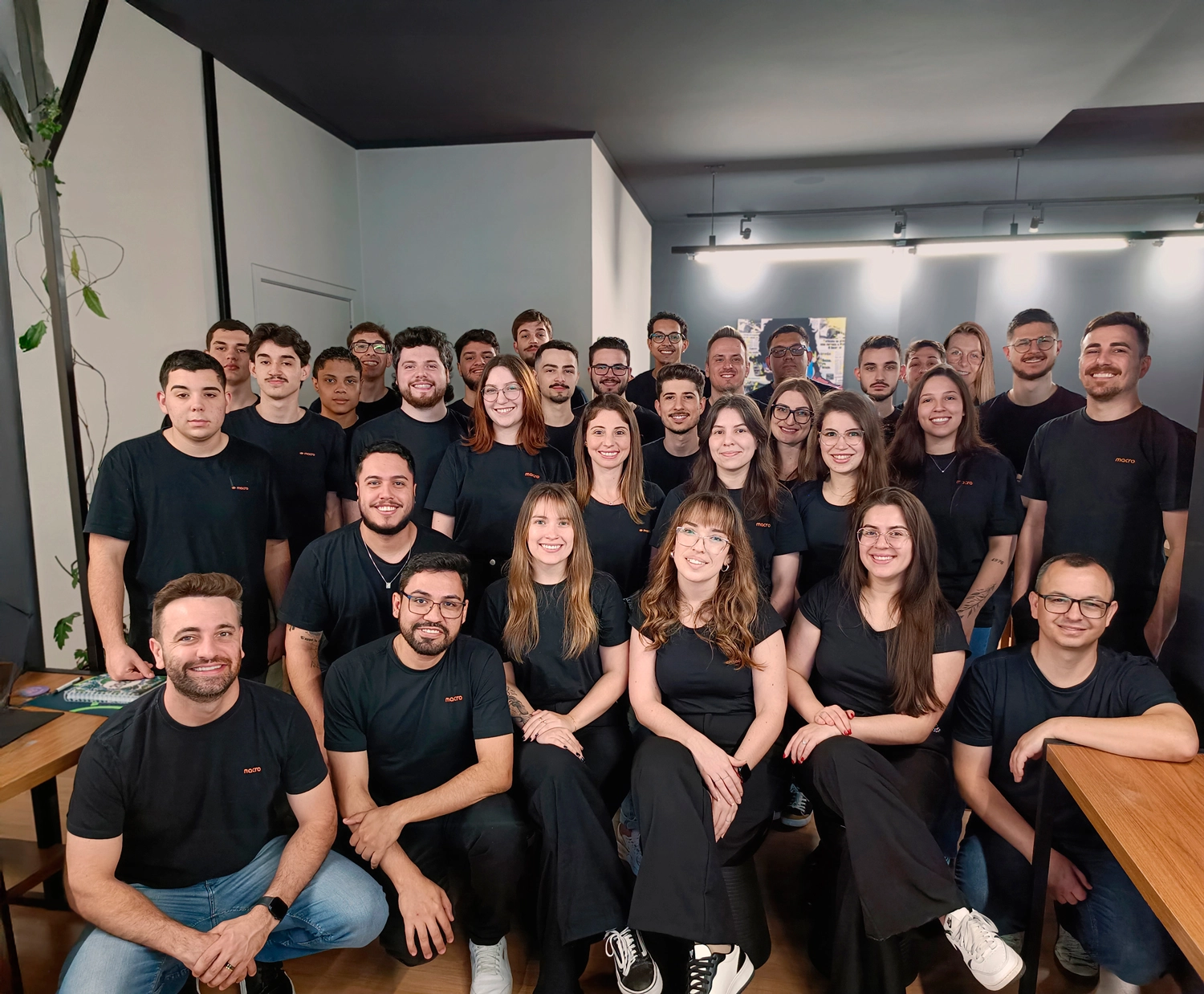 Equipe da Macro reunida para foto em grupo dentro do escritório, todos vestindo camisetas pretas.