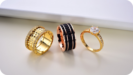 Trio de anéis — dourado, rosé e preto — exibidos lado a lado sobre superfície clara.